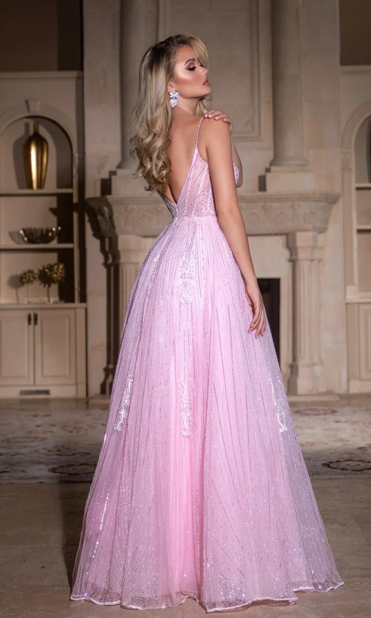 A woman stands indoors by a fireplace wearing the Portia and Scarlett PS25310 long prom dress—a floor-length, light pink, sparkly beaded gown featuring a plunging neckline and deep V-back.
